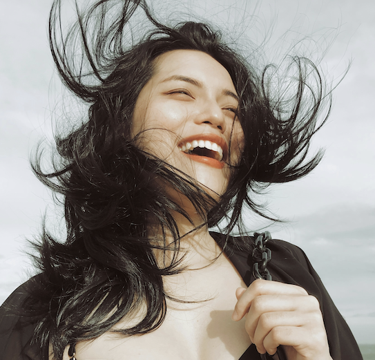 A woman with long black hair beams with joy outdoors under a cloudy sky, wearing a black top and holding the strap of her Cultivating Joy bag as the wind playfully tousles her hair.